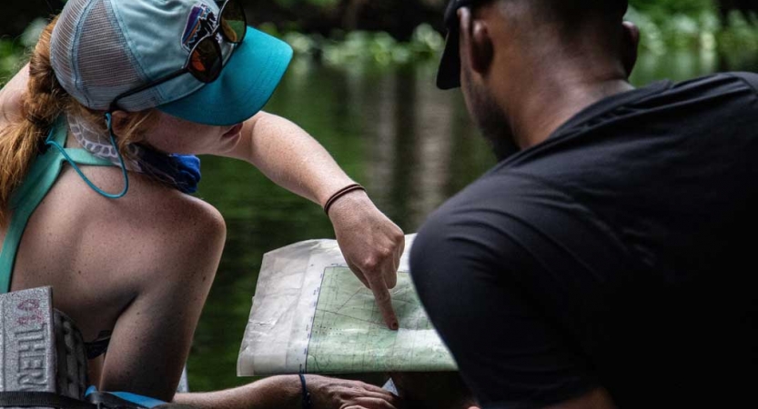 Two people who appear to be sitting in canoes examine a map. One person points at the map.